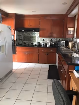 Interior view of a kitchen with wooden cabinets, a white refrigerator on the left, a black countertop with various kitchen appliances including a microwave, coffee maker, and toaster. The floor is tiled with white tiles and there is a black mat in front of the sink area.