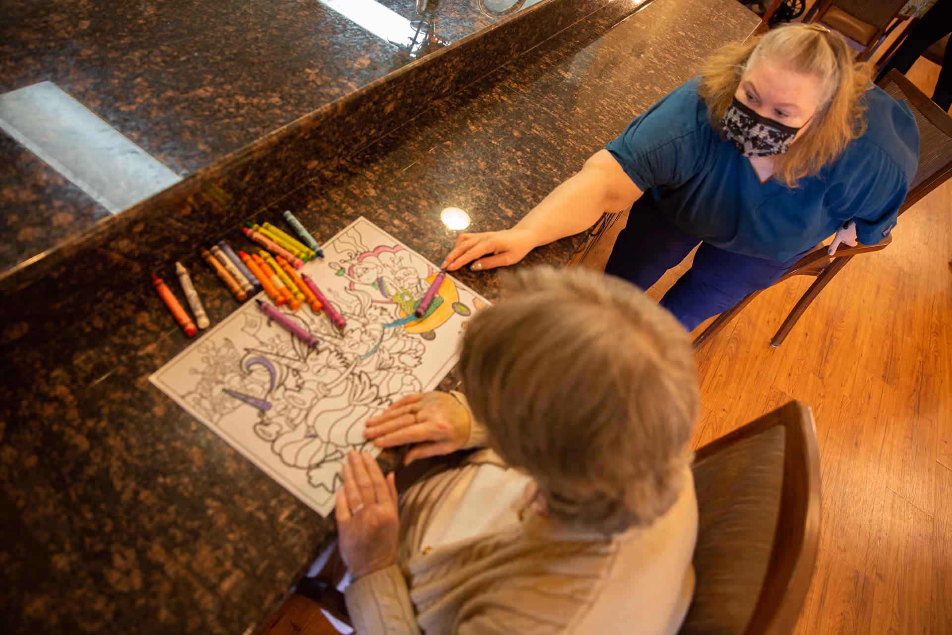 An elderly person sitting at a table coloring a detailed coloring page with crayons, while a caregiver wearing a mask leans over to assist.