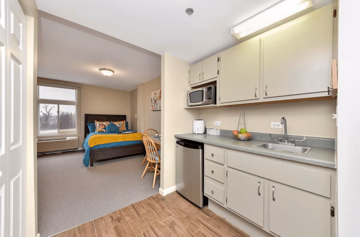View of a studio apartment in Royal Estates Assistive Living showing a small kitchen area with a sink, countertop, cabinets, microwave, and mini refrigerator. Adjacent to the kitchen is a sleeping area with a bed, pillows, a blanket, a window, and a small dining table with two wooden chairs.