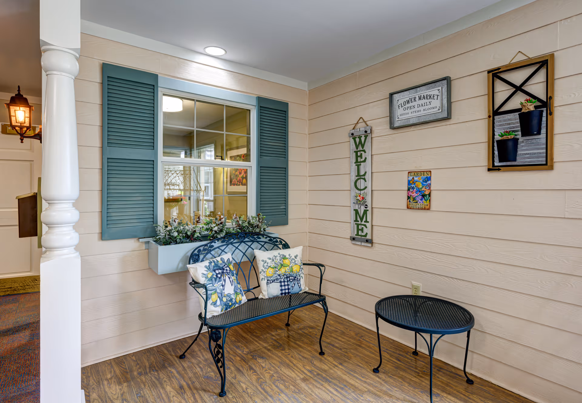 A cozy indoor seating area with a black metal bench adorned with two decorative pillows featuring floral and lemon designs. Next to the bench is a small round black metal table. The wall behind has beige horizontal paneling with decorative signs including a vertical 'WELCOME' sign, a 'FLOWER MARKET OPEN DAILY' sign, and a small colorful garden-themed plaque. A window with teal shutters and a flower box filled with greenery is also visible. The floor has a wood-like finish, and a white column is partially visible on the left side.