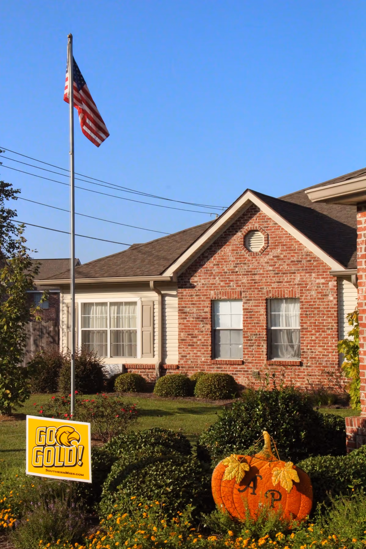 Exterior view of a brick building with a gabled roof, an American flag on a flagpole, green bushes, and a large decorative pumpkin with the letters 'AP' on it. There is also a yellow sign that reads 'GO GOLD! SouthernMiss.com' in the landscaped front yard.