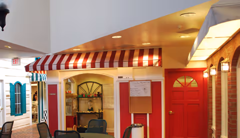 Indoor common area with café-style storefront facades, a red door and striped awning, and tables and chairs.