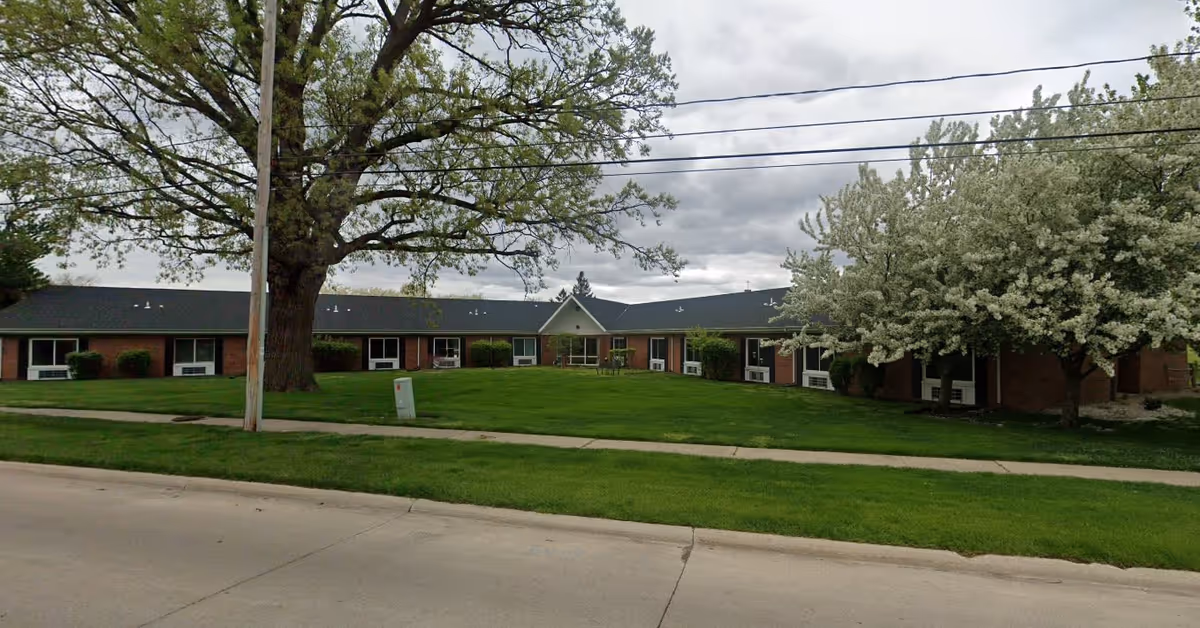 Single-story brick building with a dark roof surrounded by green grass, a large tree, and blooming white-flowered trees under a cloudy sky, viewed from across a street with a sidewalk.