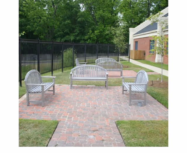 Outdoor seating area with four wooden benches arranged on a brick-paved patio surrounded by grass, next to a building with a brick exterior and a black metal fence enclosing the area. Trees and greenery are visible in the background.