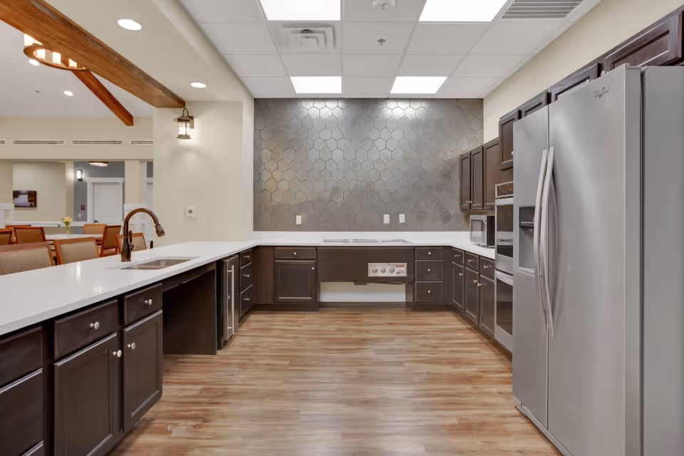 Modern kitchen area with dark wood cabinets, white countertops, a stainless steel refrigerator, built-in oven, microwave, and a sink with a bronze faucet. The backsplash features large hexagonal tiles in a muted gray tone. The kitchen opens into a dining area with wooden chairs and tables. The ceiling has recessed lighting and a wooden beam detail.