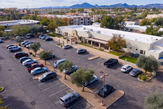 Aerial view of Shea Post Acute Rehabilitation Center showing a single-story building with a covered entrance, surrounded by a parking lot with several parked cars and trees. In the background, there are residential buildings and mountains under a clear sky.