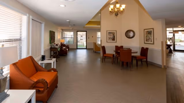 Interior view of a senior living facility common area with orange armchairs, a small round table with four chairs, framed artwork on the walls, and large windows letting in natural light.
