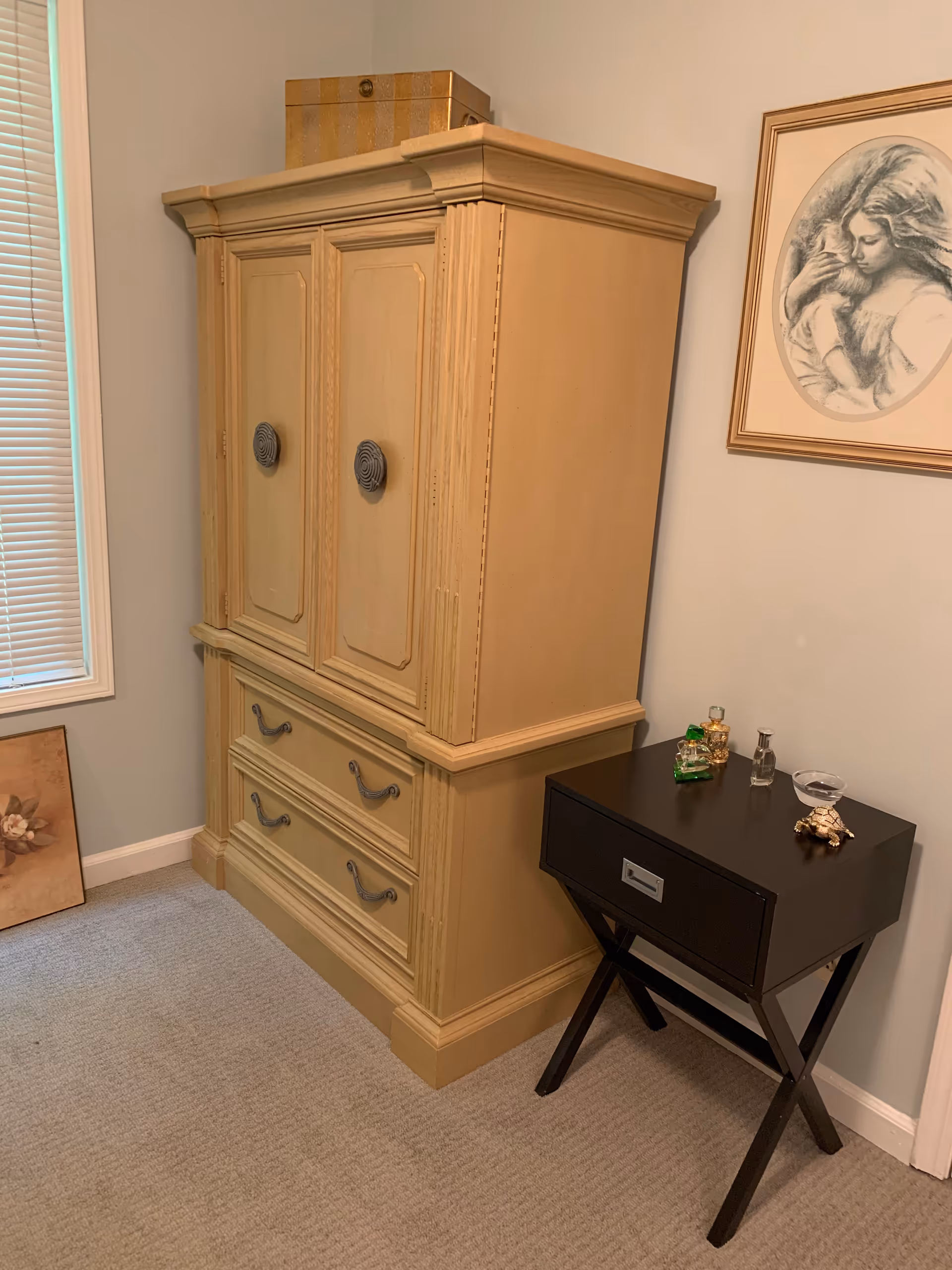 A corner of a room featuring a large beige wooden cabinet with two drawers and two doors, a small black side table with a drawer holding several small decorative items, a framed sketch of a woman holding a child on the wall, a window with closed blinds, and a framed floral picture resting on the floor.