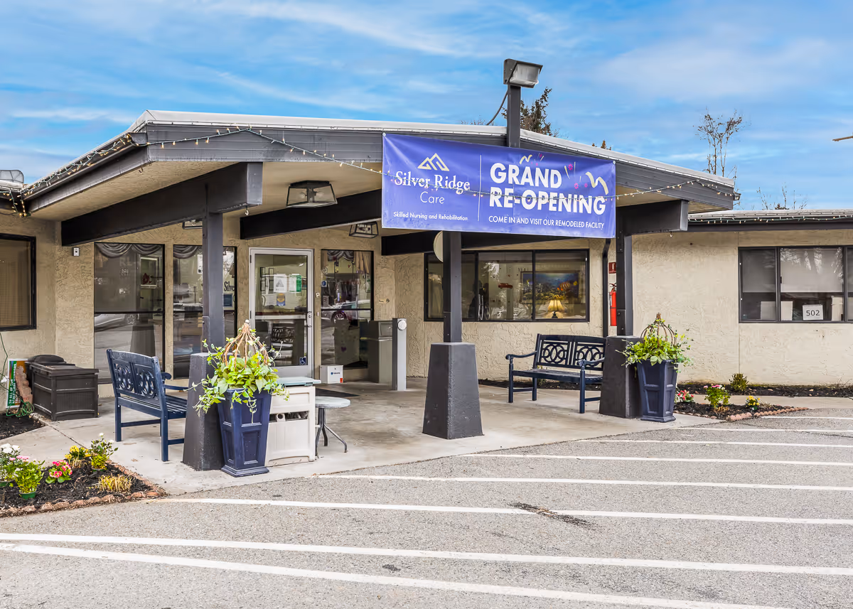 Exterior view of Silver Ridge Care facility entrance with a covered porch, benches, potted plants, and a blue banner announcing the grand re-opening of the remodeled facility.