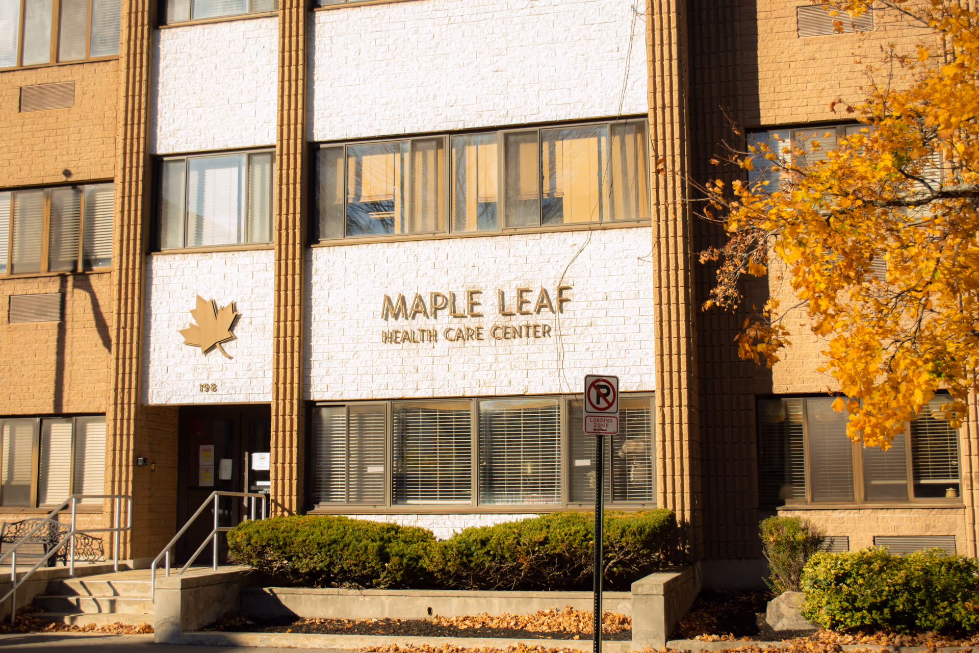 Front facade and entrance of the Maple Leaf Health Care Center building with its sign, maple leaf emblem, stairs, and an autumn tree nearby.