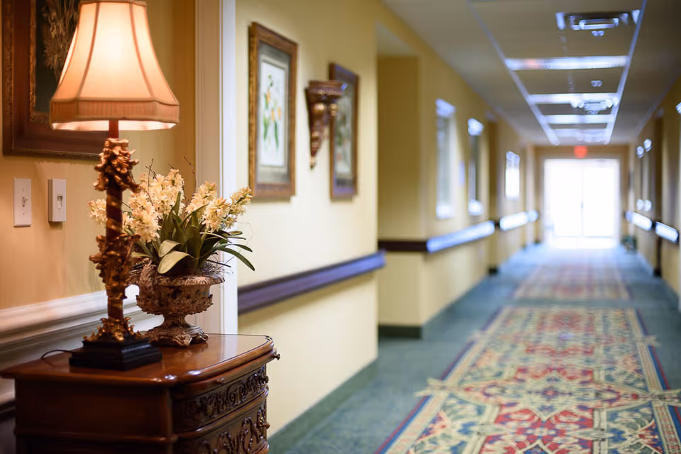 A well-lit hallway in a senior living facility with yellow walls, decorative framed artwork, and handrails on both sides. A wooden table with an ornate lamp and a flower arrangement is positioned on the left side near the foreground. The hallway has a patterned carpet and leads to a bright exit door at the far end.