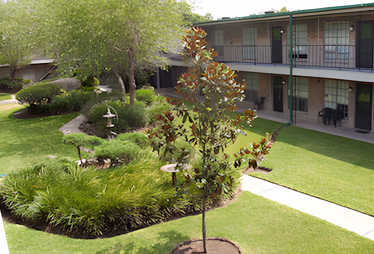 Outdoor courtyard area of Oak Shadows facility with green grass, landscaped garden beds, a small tree in the foreground, and a two-story building with balconies and chairs in the background.