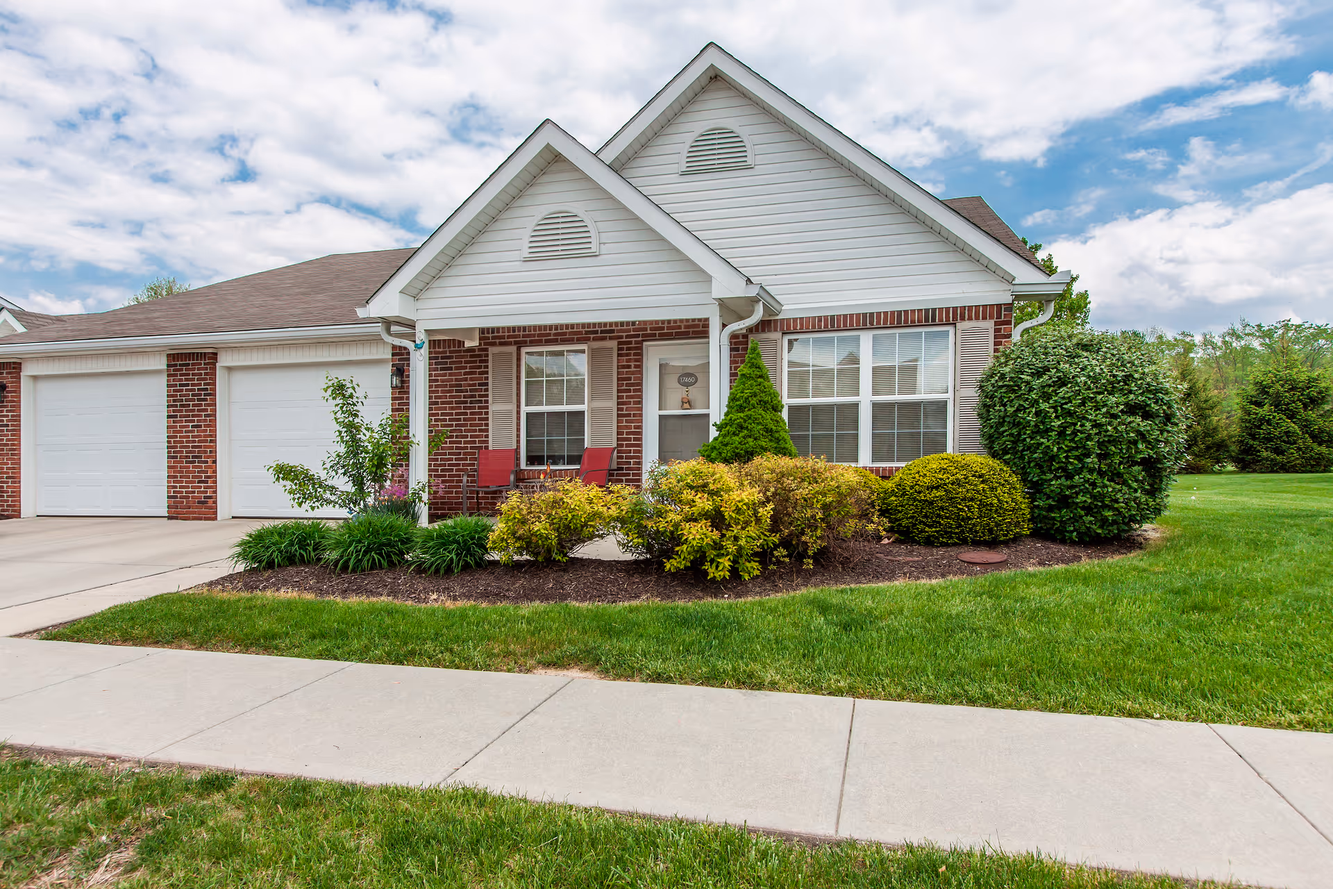 Front exterior of a single-story brick and white-siding residence with a two-car garage, a small porch with chairs, manicured shrubs and a sidewalk.
