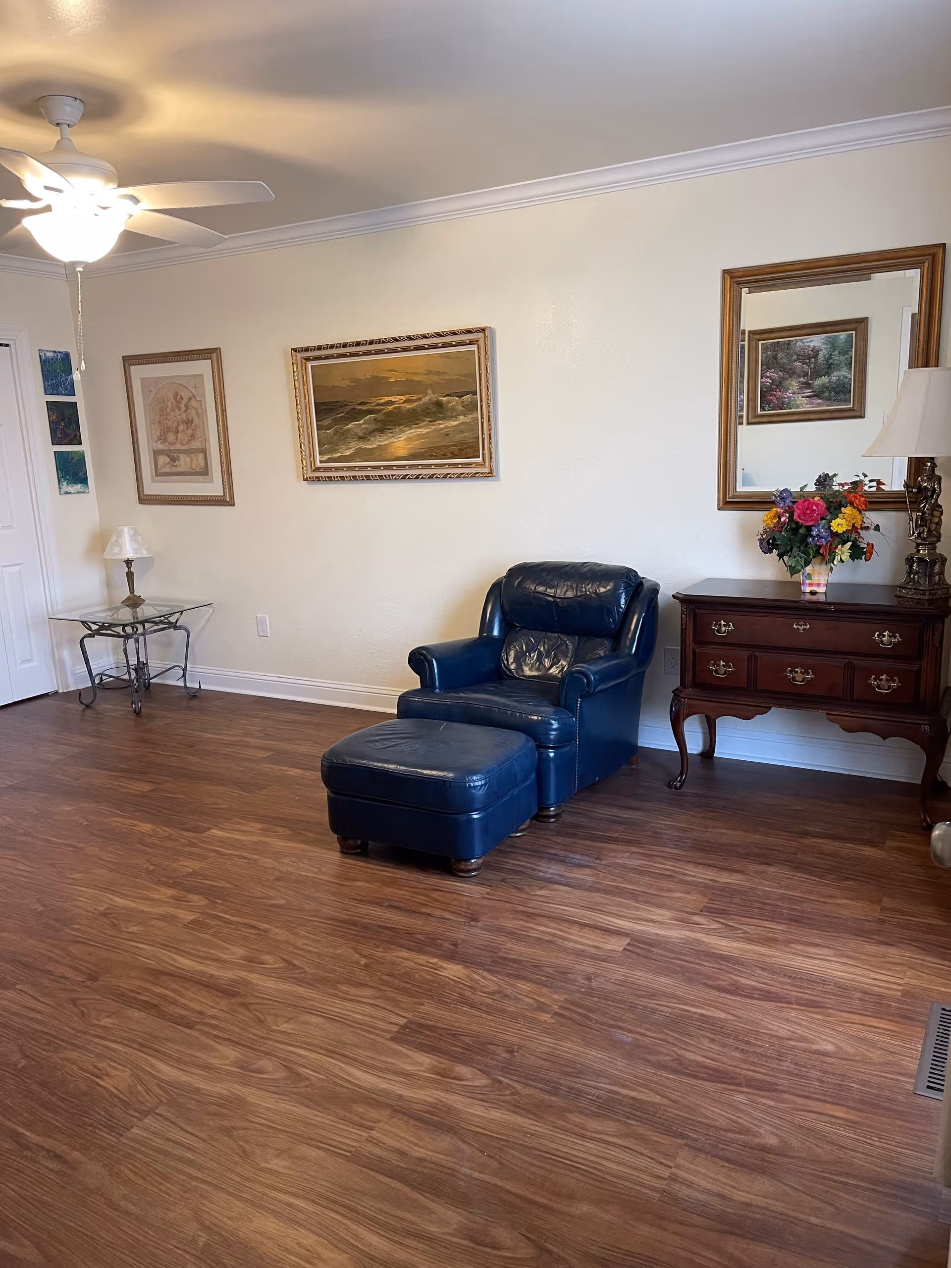 A living room with a blue leather armchair and ottoman, wooden side table and dresser with a lamp and flowers, and framed paintings on the wall.