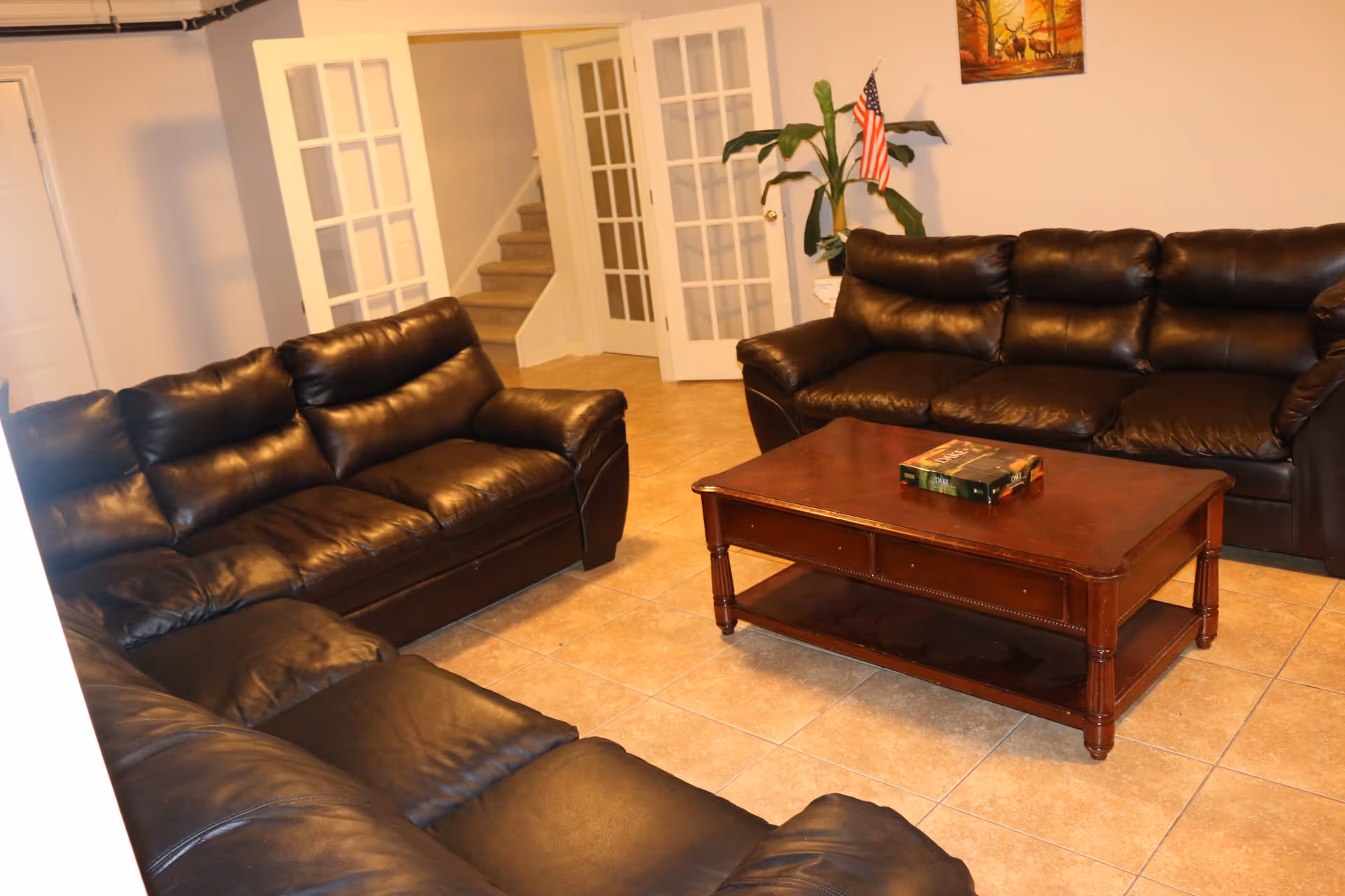 Basement living room with three dark leather sofas arranged around a wooden coffee table, with French doors and a staircase in the background.