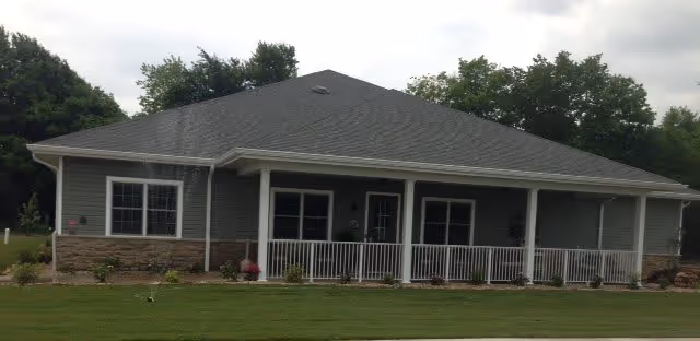 Single-story building with a gray roof and light gray siding, featuring a covered front porch with white railings. The building is surrounded by a well-maintained lawn and some small shrubs, with trees in the background under a cloudy sky.
