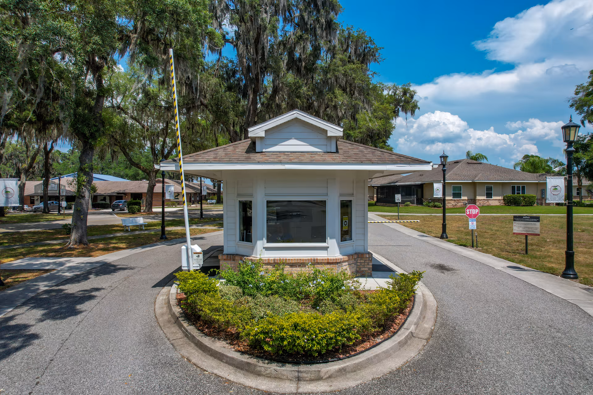 Small white security or gatehouse building with a brown shingled roof situated in the middle of a driveway entrance, surrounded by greenery and trees with a clear blue sky above. There are stop signs, street lamps, and residential buildings visible in the background.