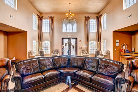 Sunlit assisted-living common room featuring a large curved leather sectional, armchairs, tall arched windows, and a central chandelier over the entrance.