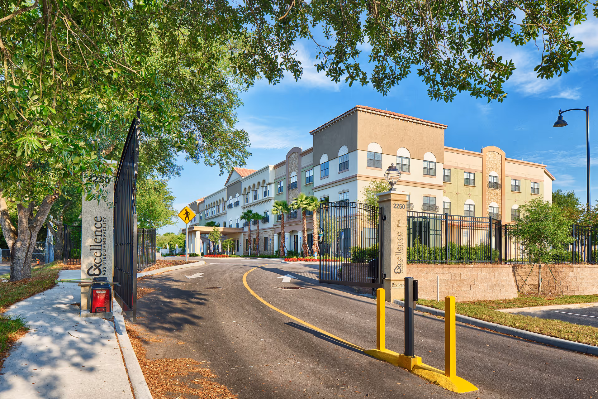 Entrance driveway to Excellence Senior Living facility with a gated entrance, trees lining the sidewalk, and a multi-story building with balconies and arched windows in the background under a clear blue sky.