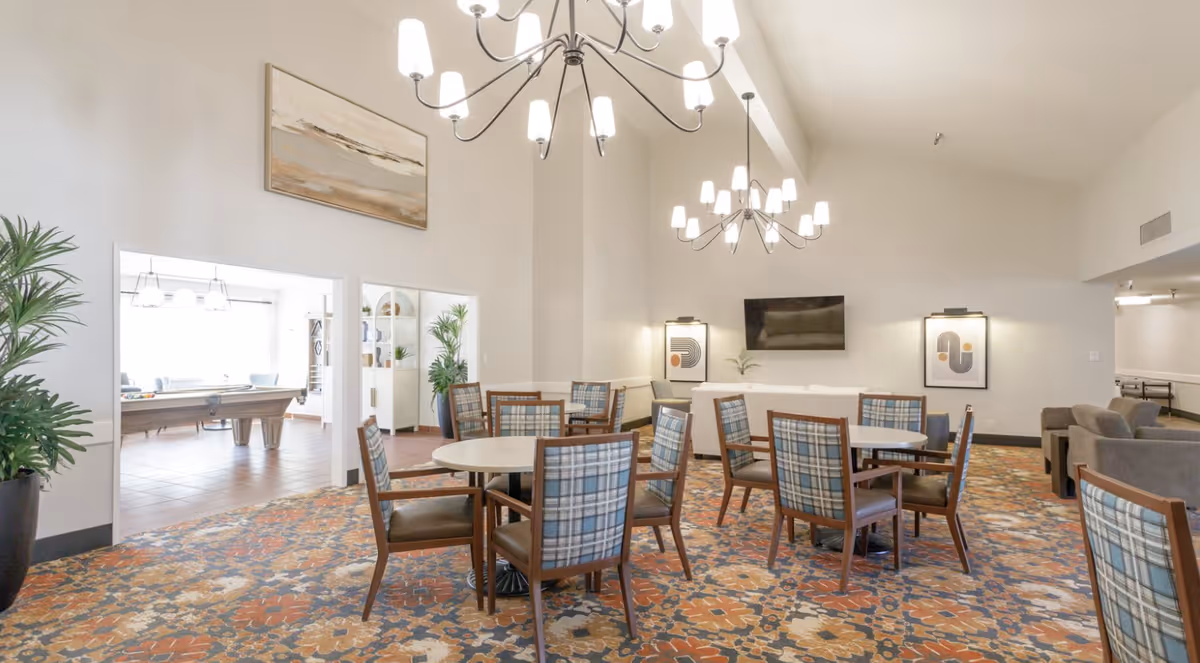 A spacious and well-lit common area with round tables and chairs arranged on a patterned carpet. The room features high ceilings with modern chandeliers, wall art, and a flat-screen TV mounted on the wall. Adjacent to this area is another room with a pool table and additional seating, visible through open doorways. There are also potted plants adding greenery to the space.