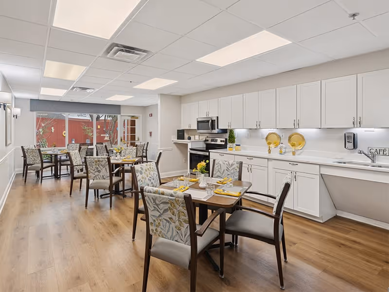 A bright dining room with wooden floors and several tables set with yellow napkins and glassware. The room features floral upholstered chairs and a kitchen area with white cabinets, a microwave, stove, and sink. Large windows at the far end let in natural light and show a glimpse of an outdoor area.