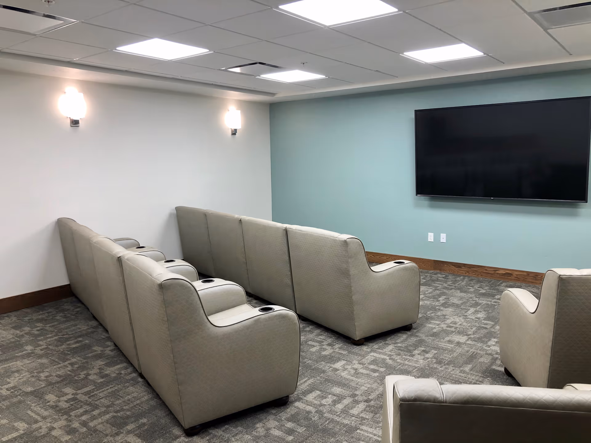 A small theater-style room with two rows of beige reclining chairs facing a large flat-screen TV mounted on a light blue wall. The room has gray patterned carpet, white walls with two wall-mounted lights, and a drop ceiling with fluorescent lighting.