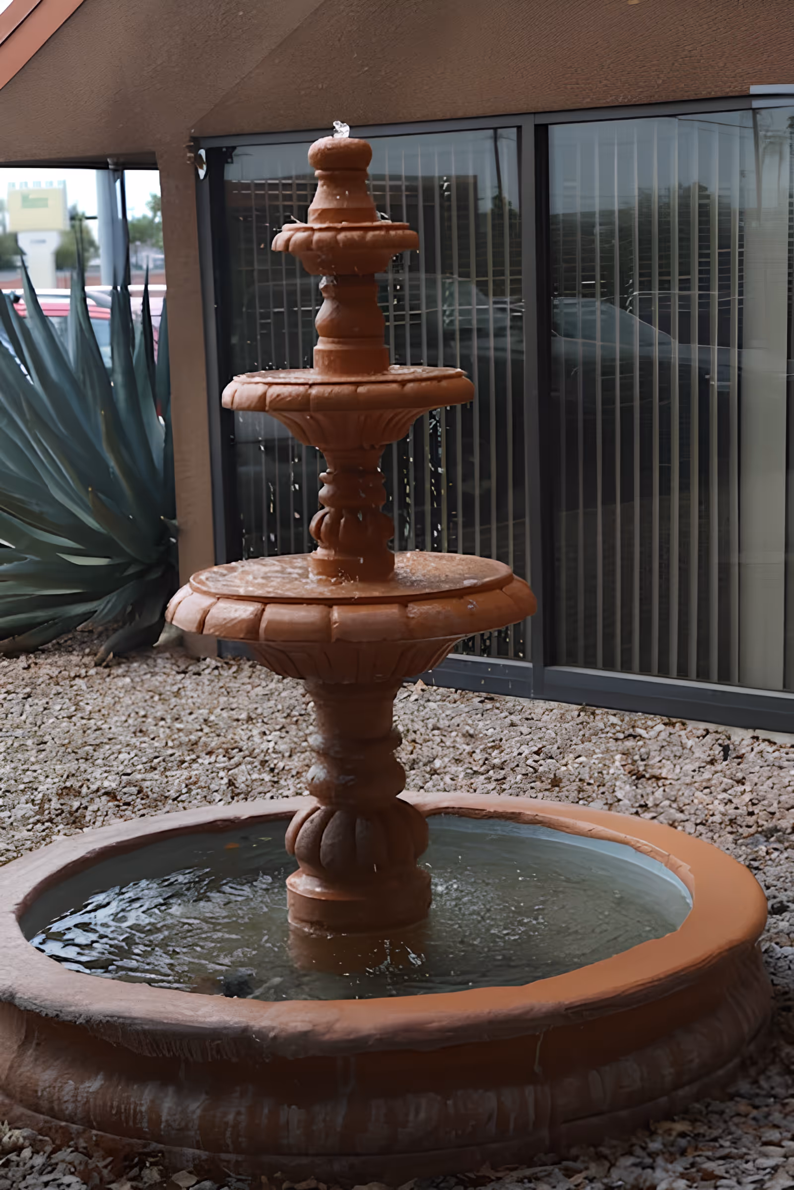 A three-tiered terracotta water fountain with water flowing from the top tier into the lower basins, surrounded by gravel and positioned in front of a building with large windows and vertical blinds.