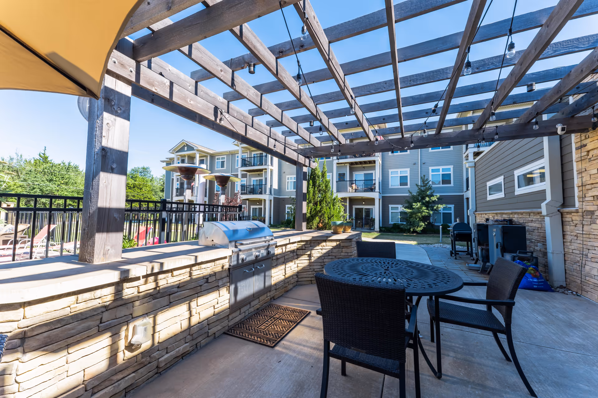 Outdoor patio area at The Cedars at Woodland Hills featuring a pergola with string lights, a built-in stone grill, a round metal table with chairs, and a view of the residential building and greenery in the background.