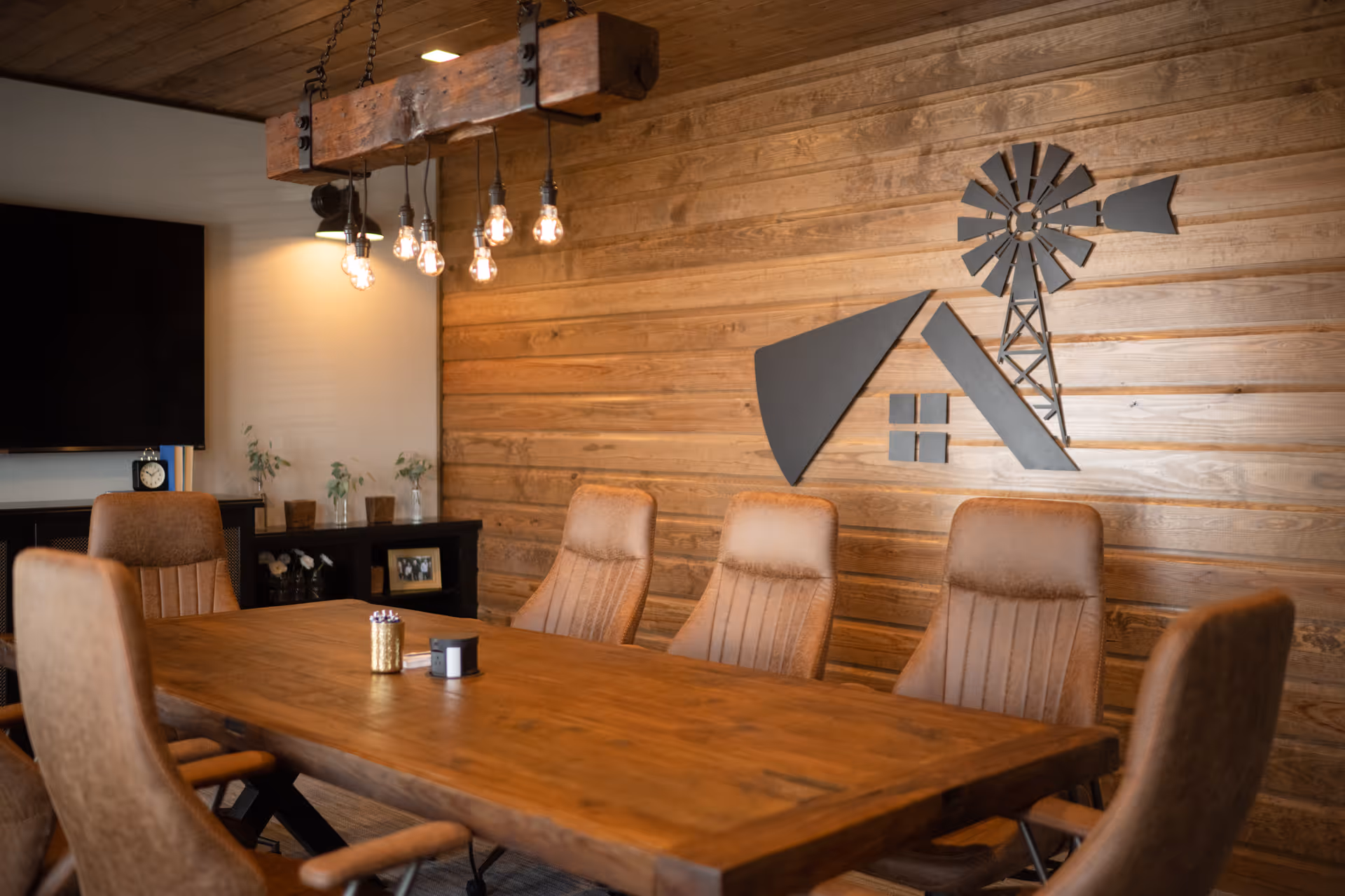 Wood-paneled interior with a long wooden table, upholstered chairs, hanging exposed-bulb light fixture and windmill wall art.
