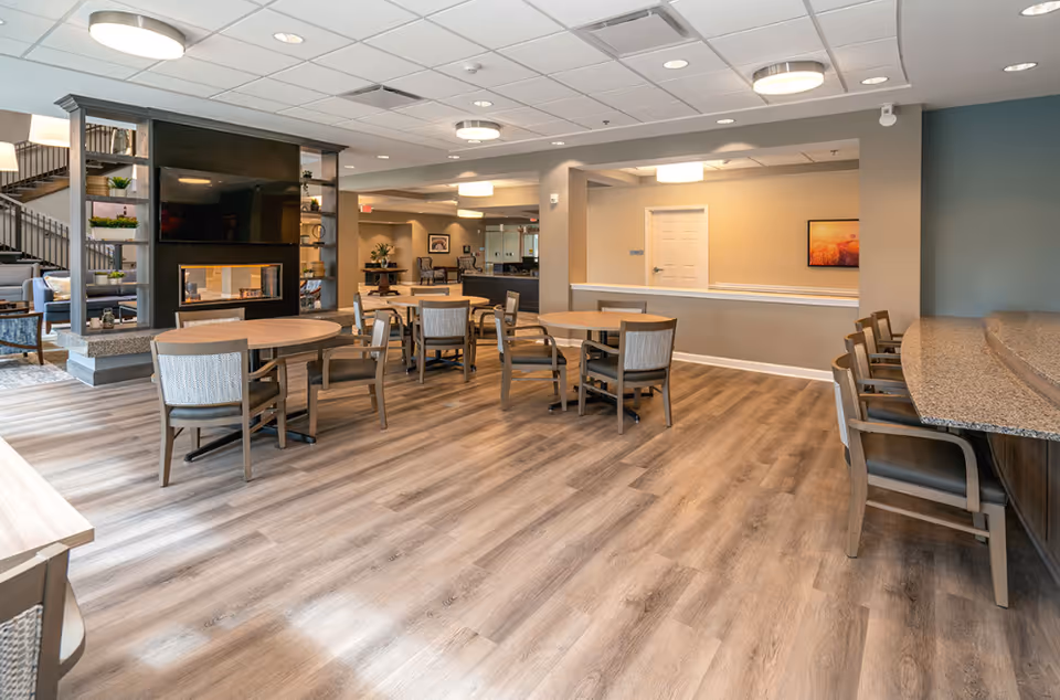 A spacious and well-lit common area in a senior living facility featuring multiple round tables with chairs, a modern fireplace with a mounted TV above it, and a seating area with armchairs. The room has wood-style flooring, recessed lighting, and a neutral color palette with beige and gray tones.