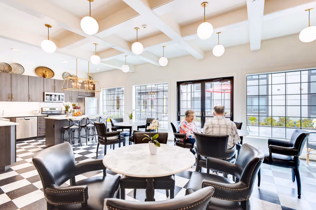 Bright and modern dining area in an assisted living community featuring black and white checkered flooring, round marble tables with black leather chairs, a kitchen area with stainless steel appliances, and large windows letting in natural light. Two elderly people are seated at a table near the windows, engaged in conversation.