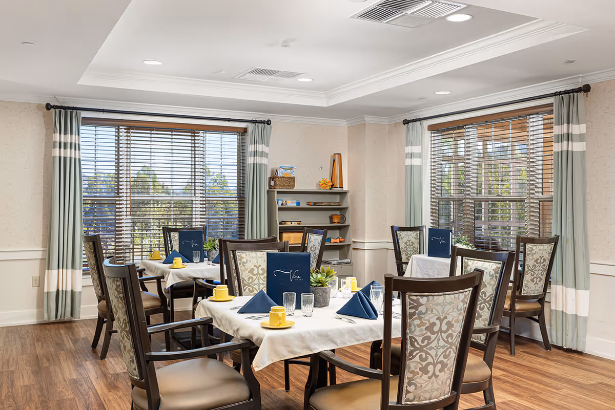 A bright dining room with wooden floors and large windows covered with wooden blinds and green curtains with white stripes. Several tables are set with white tablecloths, blue napkins, yellow cups and saucers, and clear drinking glasses. The chairs have patterned upholstery on the backrests. A small shelf with decorative items is against the wall.