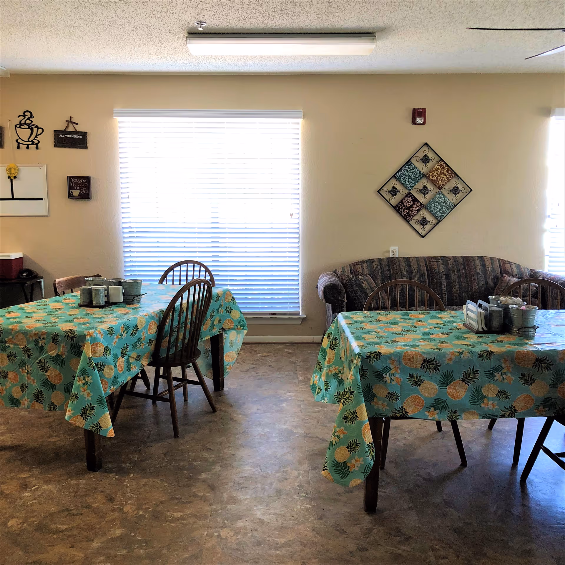 Communal dining area with two tables covered in pineapple-patterned tablecloths, wooden chairs, and a couch in front of a window.