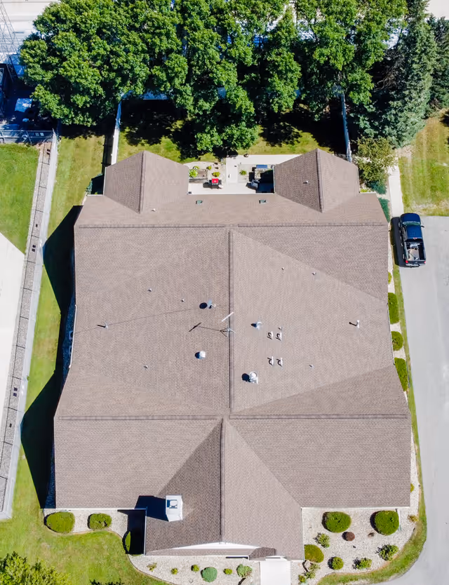 Aerial view of a senior living facility building with a brown roof, surrounded by green lawns, trees, and a driveway with a parked vehicle on the right side.