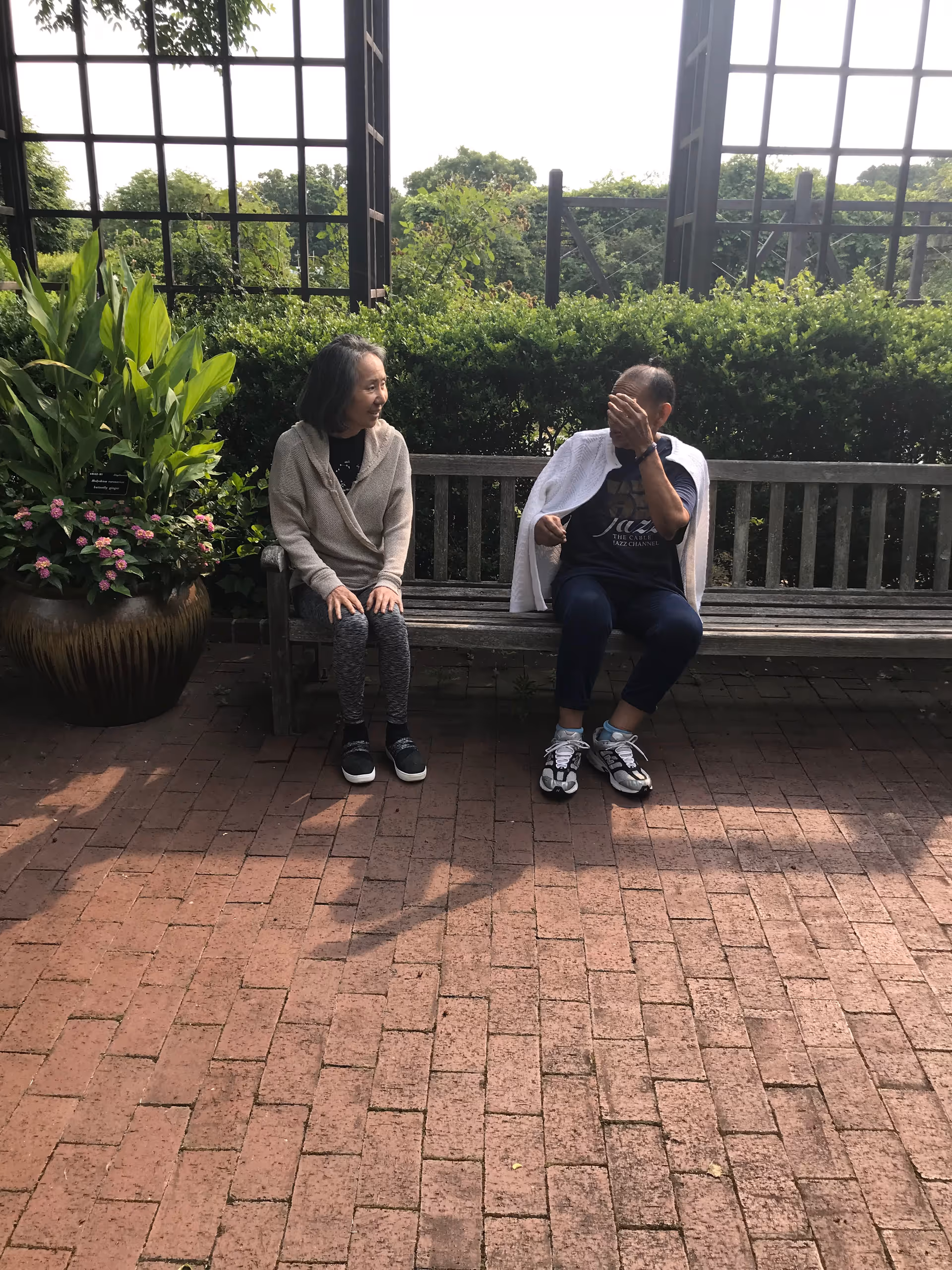 Two older adults sit on a wooden bench in a garden courtyard, with one person covering their face.