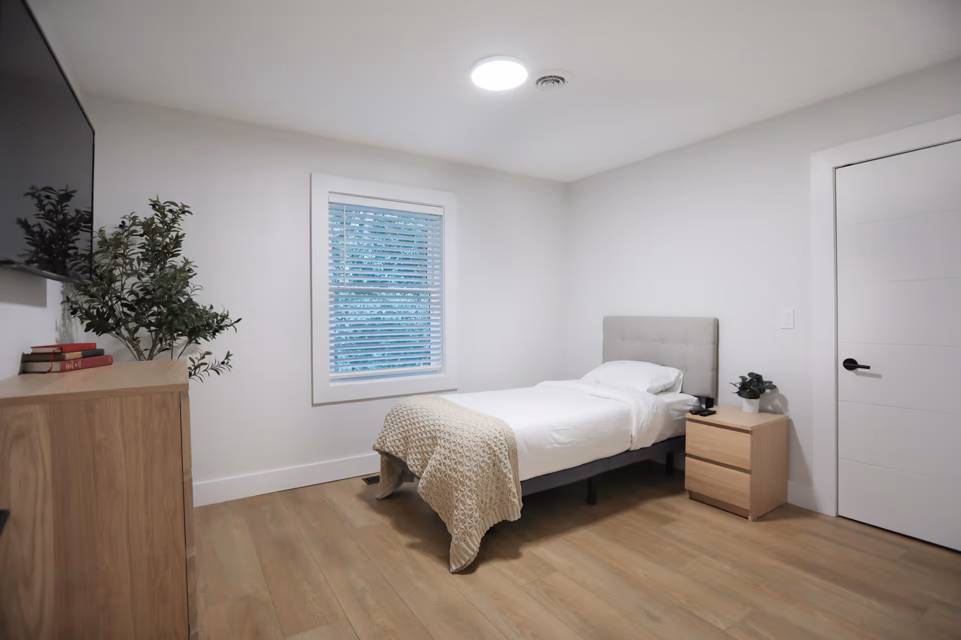 A simple, clean bedroom with a single bed featuring a tufted headboard, white bedding, and a beige knitted throw. There is a wooden nightstand with a small potted plant next to the bed, a wooden dresser with books and a plant on top, a window with white blinds, and a wall-mounted TV. The room has light-colored wooden flooring and white walls.