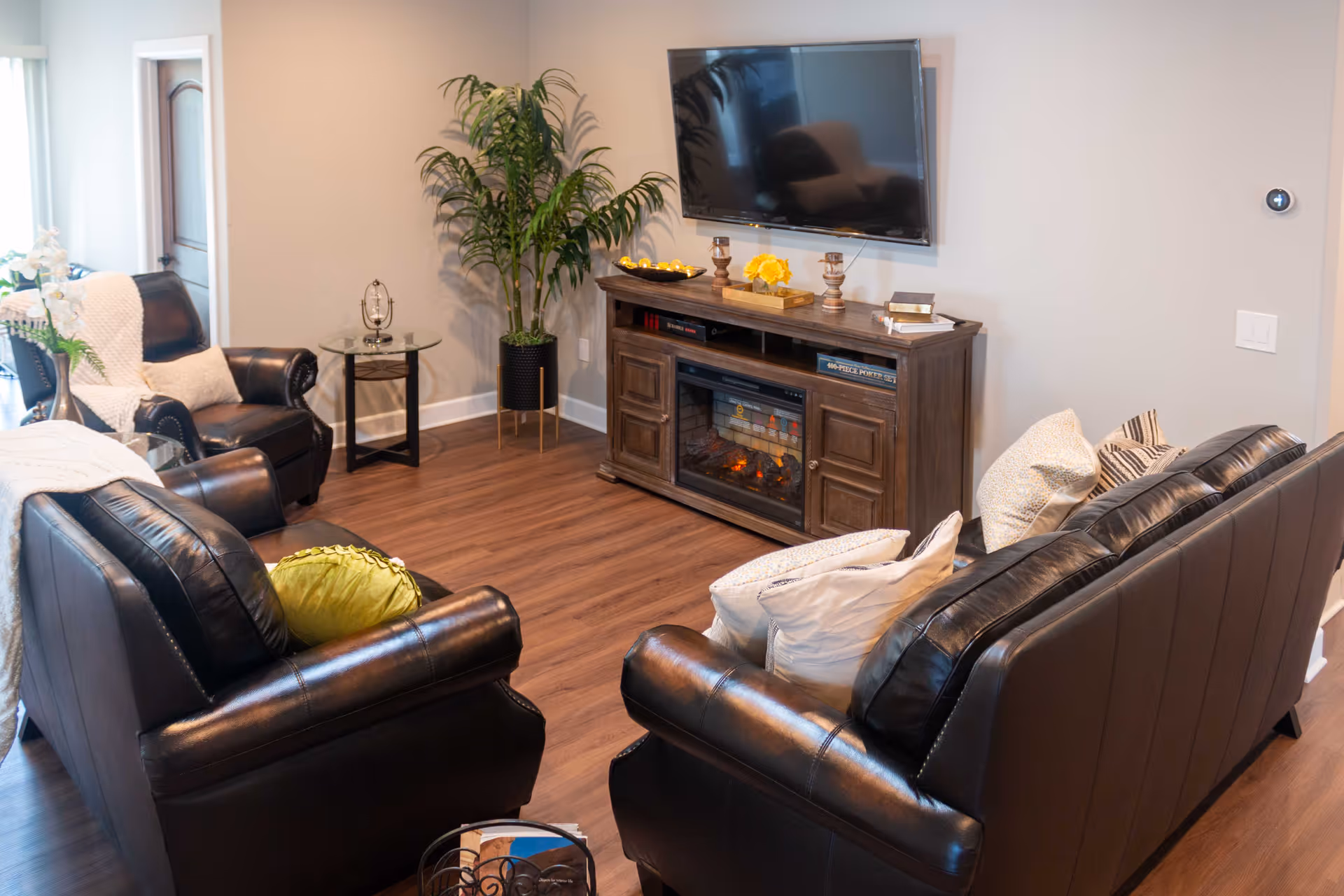 A cozy living room with dark leather sofas and armchairs arranged around a wooden entertainment center with an electric fireplace. A flat-screen TV is mounted on the wall above the fireplace. There is a tall green plant in a black pot in the corner, a small round side table with a decorative item, and several pillows on the sofas. The room has wooden flooring and light-colored walls.
