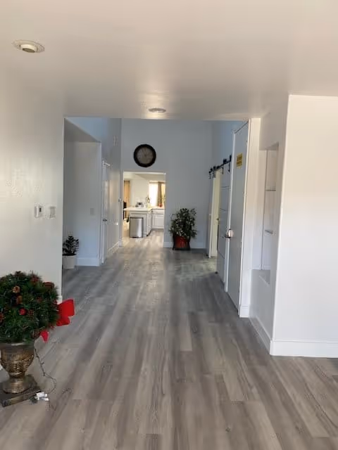 A long hallway with light wood flooring and white walls leading to a kitchen area in the background. There are two potted plants on either side of the hallway, a clock on the far wall, and a sliding barn door on the right side. The space is well-lit with recessed ceiling lights.