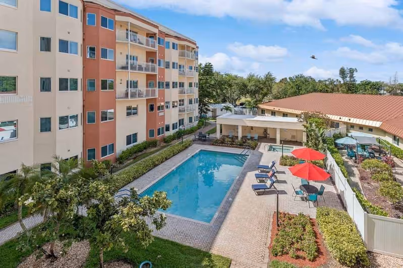 Outdoor view of a senior living facility featuring a rectangular swimming pool surrounded by lounge chairs and tables with red umbrellas. The pool area is adjacent to a multi-story building with balconies and windows. There are landscaped bushes and trees around the pool area under a partly cloudy sky.