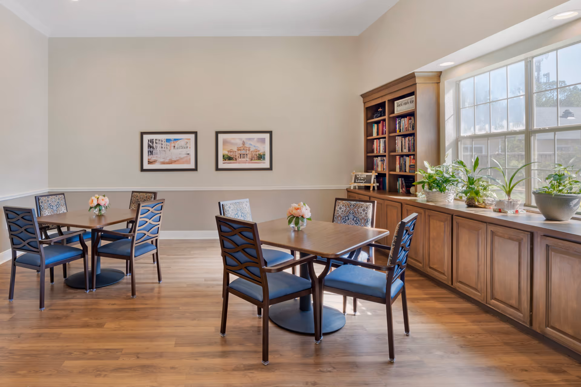 A bright and cozy senior living facility room with two wooden tables, each surrounded by four chairs with blue cushions. The room features a large window letting in natural light, a wooden bookshelf filled with books, and several potted plants on a wooden cabinet beneath the window. Two framed pictures hang on the beige wall.