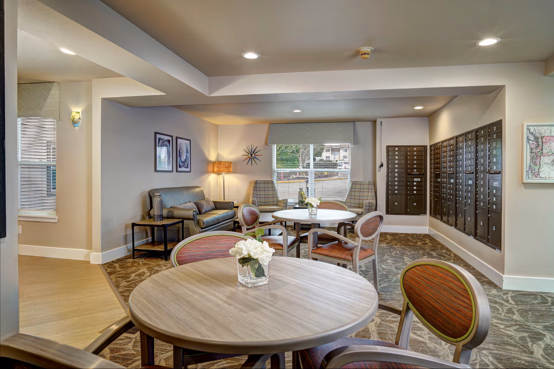 Interior common area of a senior living facility with round tables and chairs, a seating area with a sofa and two armchairs, a floor lamp, wall art, and a wall of mailboxes. A window with a valance lets in natural light.