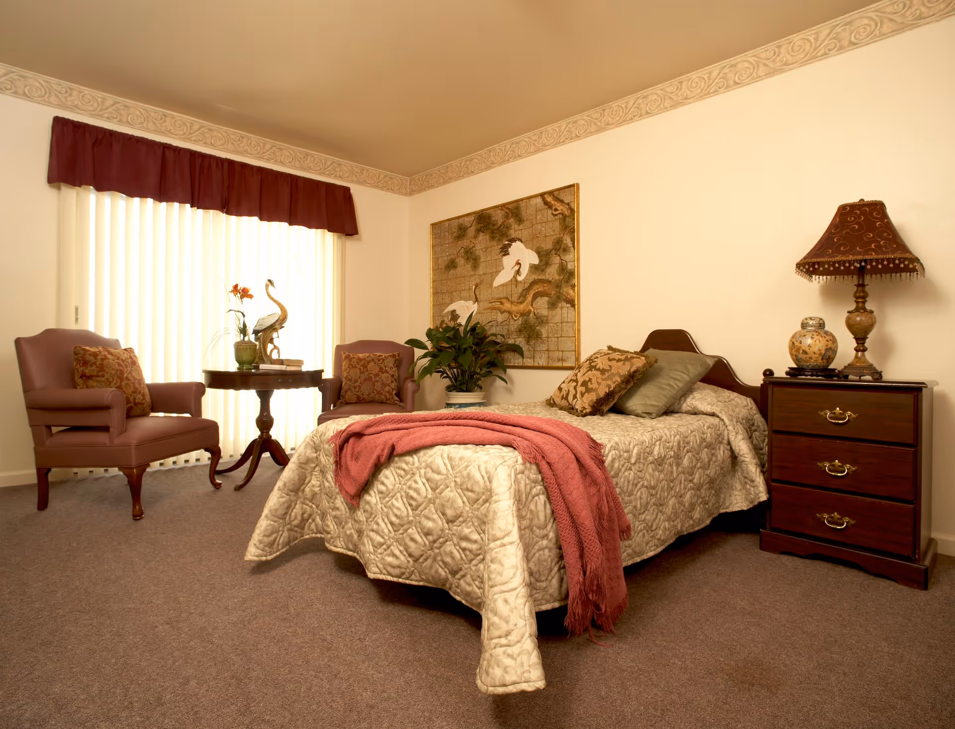 A cozy bedroom in a senior living facility featuring a bed with a beige quilted bedspread and a red throw blanket. There are two pillows on the bed. To the right of the bed is a wooden nightstand with three drawers, a decorative lamp, and a vase. On the left side of the room, there are two upholstered armchairs with patterned cushions and a round wooden table between them, holding a decorative bird sculpture and a small plant. A large window with vertical blinds and a red valance lets in natural light. A framed artwork depicting birds hangs on the wall above a potted plant.