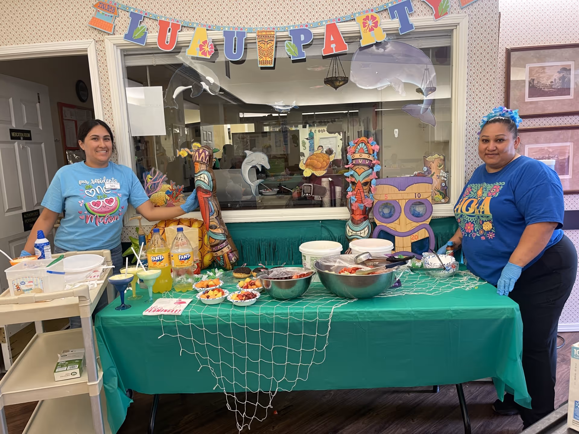Two women standing behind a table decorated for a luau party with tropical-themed decorations including tiki statues, dolphins, and a banner that reads 'LUAU PARTY'. The table is covered with a green tablecloth and has bowls of fruit, bottles of Fanta soda, and other food items. The women are smiling and wearing casual clothing, one in a light blue shirt and the other in a dark blue shirt with a floral headband.