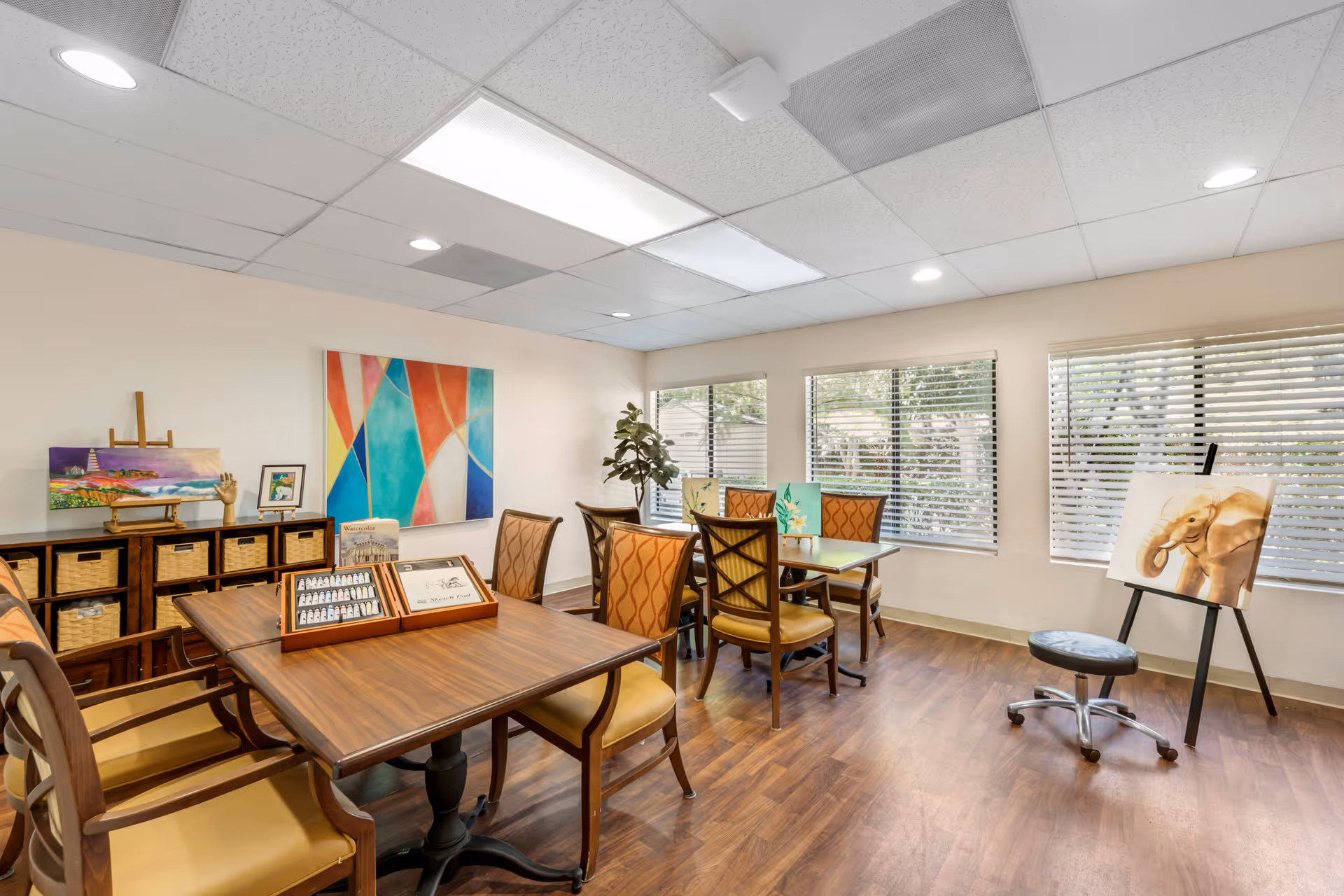 A bright room with wooden floors and multiple tables and chairs arranged for group activities. The room has large windows with blinds letting in natural light. There are paintings displayed on easels and a colorful abstract painting on the wall. A storage unit with baskets and art supplies is visible along one wall.