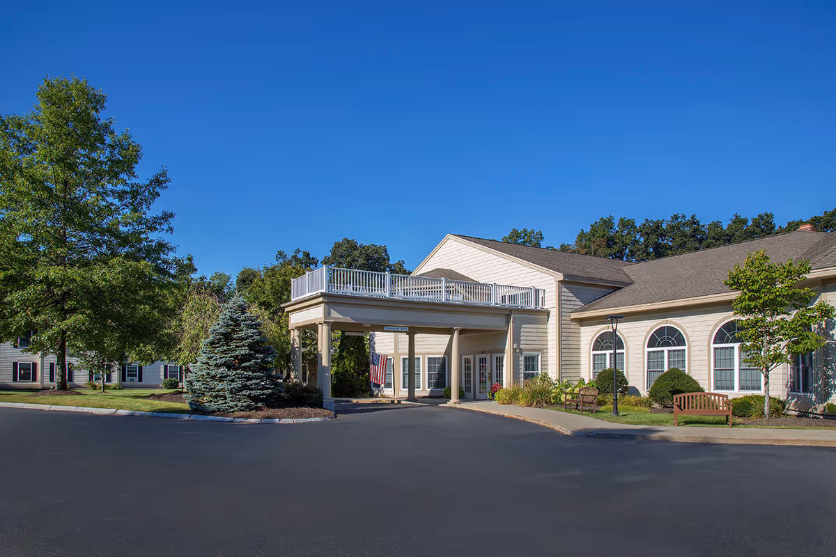 Exterior view of Benchmark Senior Living at Shrewsbury Crossings showing a beige building with a covered entrance, surrounded by trees and landscaping under a clear blue sky.