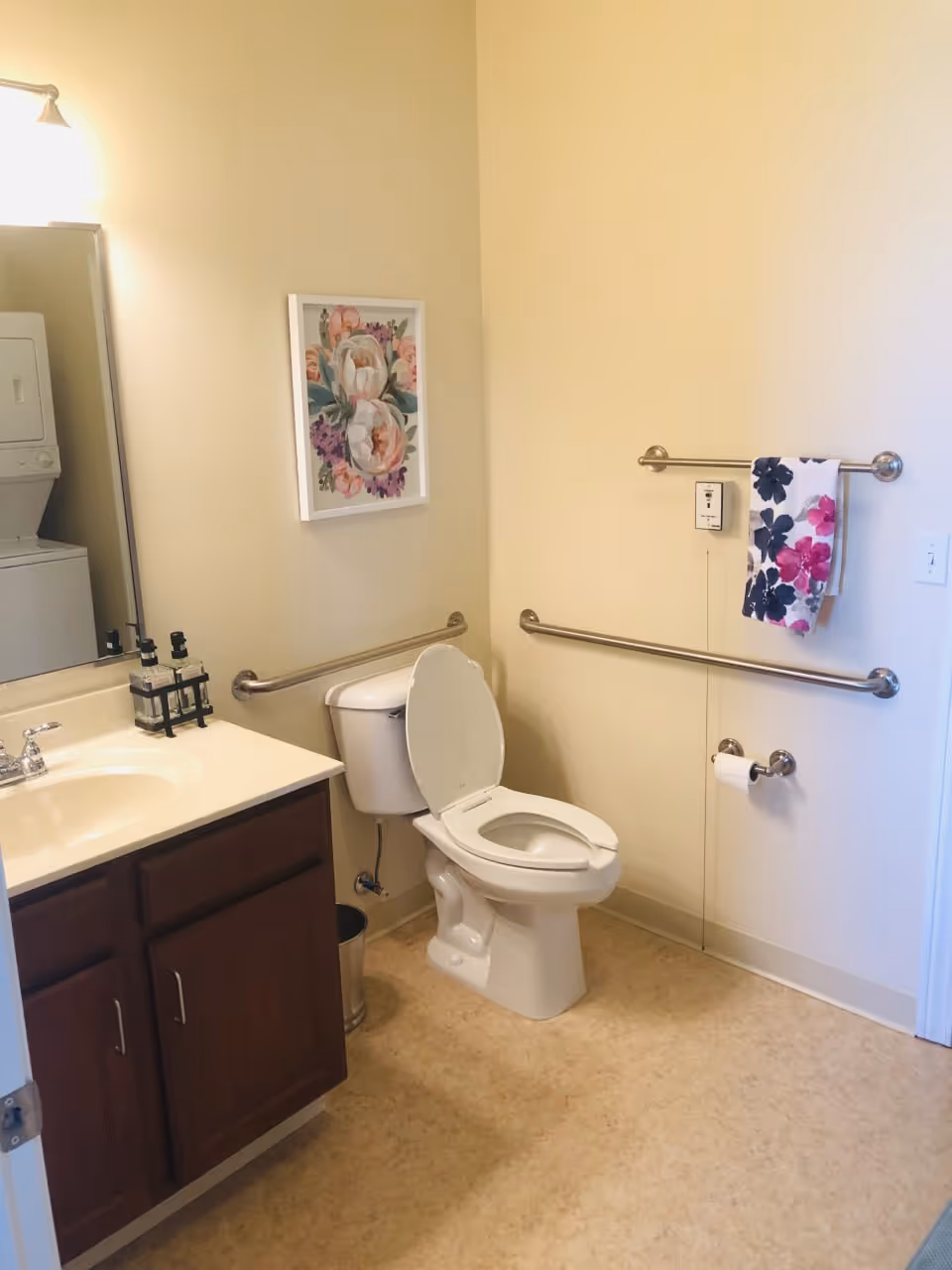 A clean bathroom with a white toilet with the lid open, a wooden cabinet with a sink and faucet, a large mirror, and a floral painting on the wall. There are grab bars on the walls near the toilet, a towel rack with a floral towel, and a toilet paper holder. A stacked washer and dryer unit is partially visible in the mirror reflection.