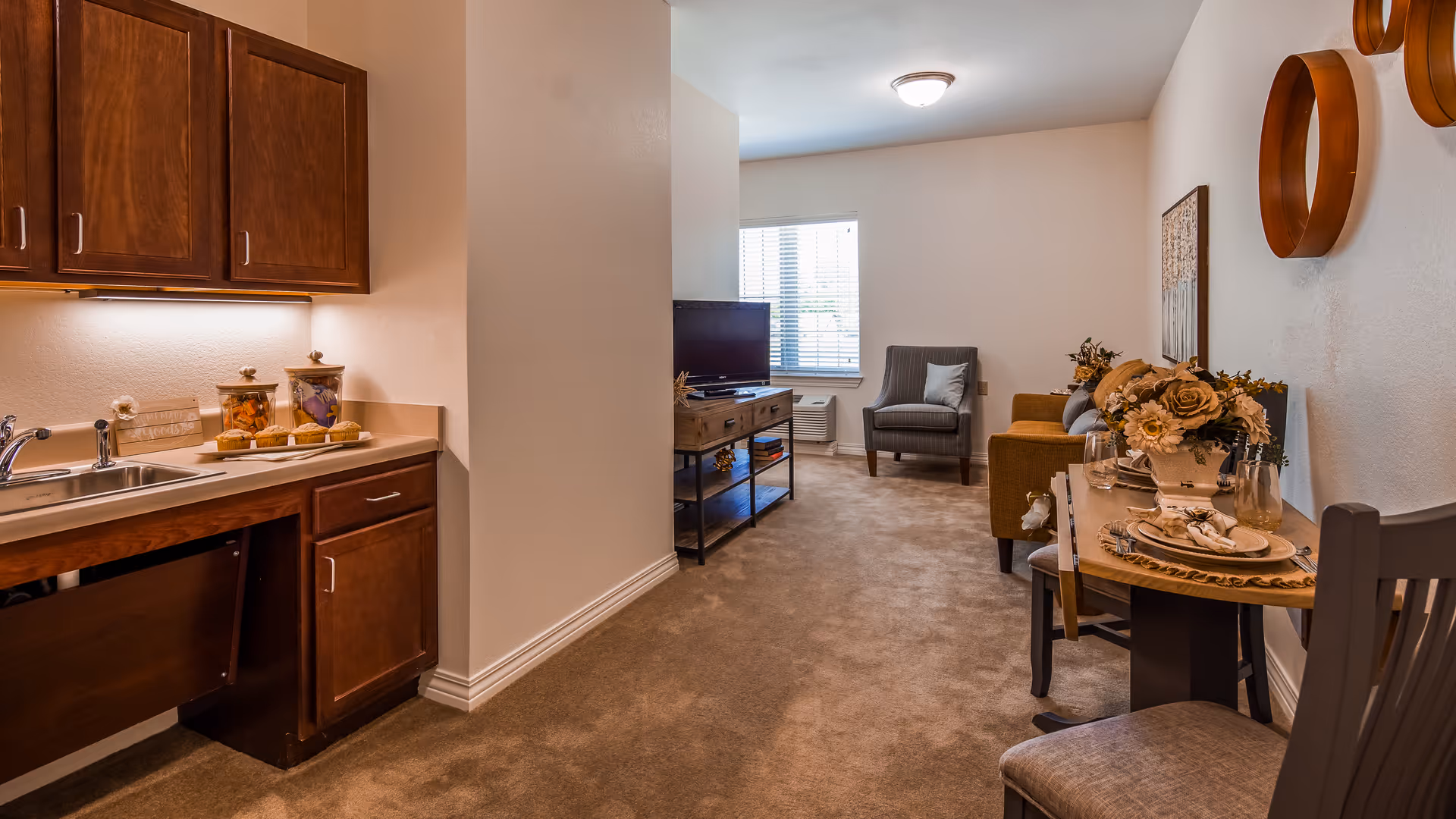 Interior view of a senior living facility unit showing a small kitchenette with wooden cabinets and a sink on the left, leading into a living area with a TV on a stand, an armchair, a sofa, and a small dining table set for two with floral centerpiece and wall decorations.