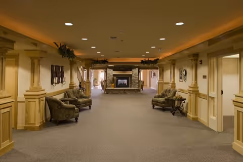 A wide hallway in an assisted living facility with beige walls and carpeted floor. The hallway features decorative columns and trim, with comfortable upholstered chairs and small tables along the sides. At the far end, there is a stone fireplace with a fire burning, and some greenery decorations above the columns near the ceiling.