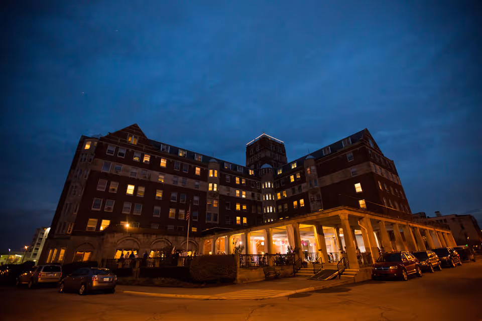The Whitcomb Senior Living Tower building at dusk with lights on inside and several cars parked outside. The multi-story brick building has a prominent central tower and a covered entrance area with columns.
