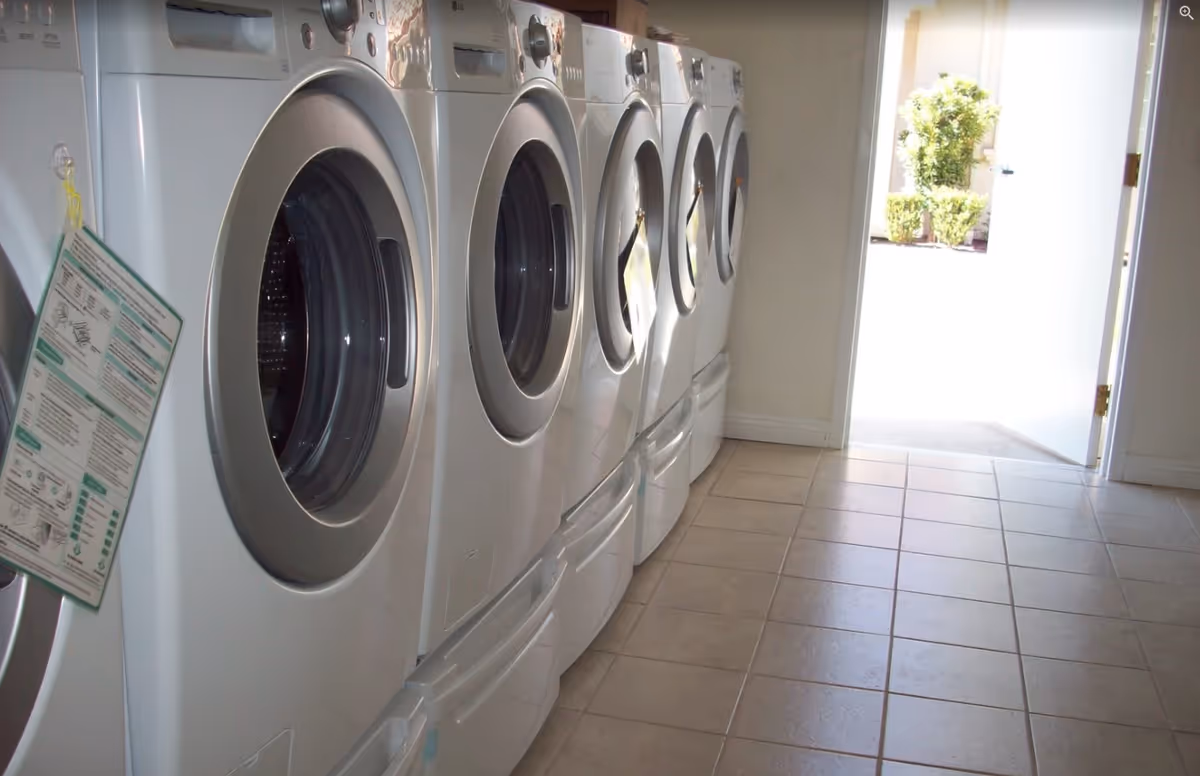 A row of front-loading washing machines in a laundry room with tiled flooring and an open door leading outside, showing some greenery.